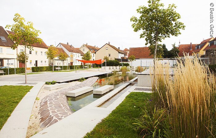 Sonnenuhrenpark Ein Park mit einem Teich. Im Teich sind kreisförmig Steinblöcke angeordnet. Sie bidlen eine Sonnenuhr. Vor dem Teich eine keline Bühne. Im Hintergund Wohnhäuser.