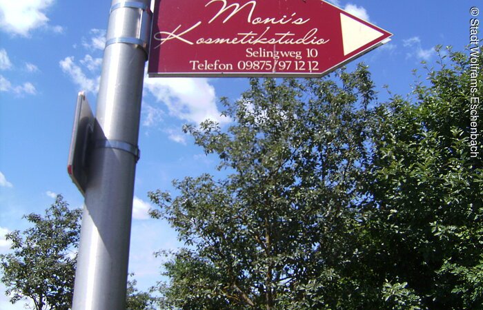 Monis Kosmetikstudio Roter Wegweiser mit Anschrift befestigt an einem Laternenmast. Darauf steht Monis Kosmetikstudio Selingweg 12 Telefon 09875 971212