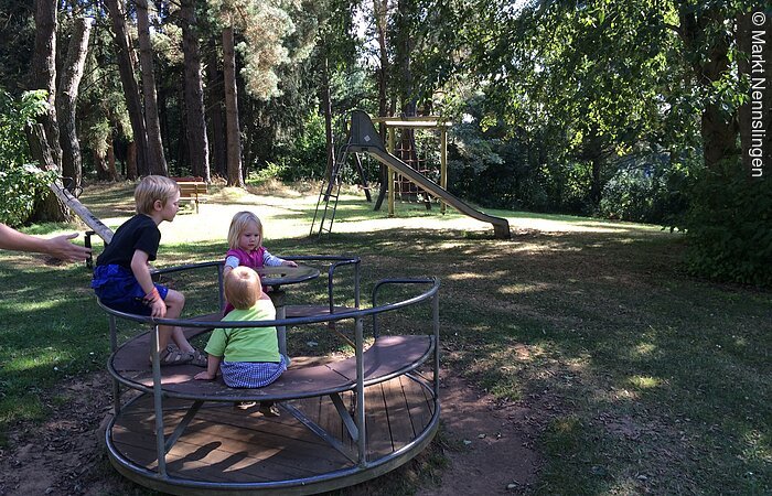 Geschwister sitzen im Rondell, der Spielplatz ist im Wald und im Hintergrund steht eine Rutsche.