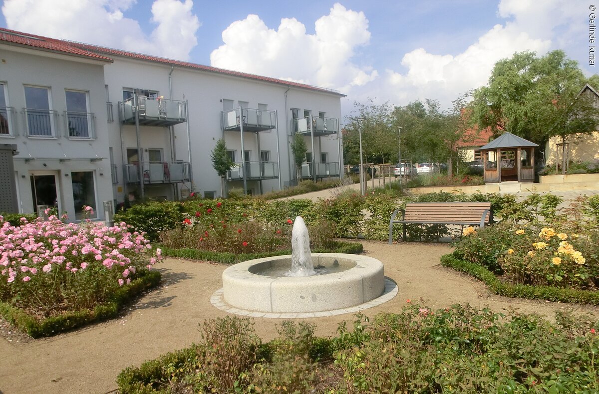 Barockgarten am Steingruberhaus in Merkendorf Ein kleiner Park mit Schotterwegen und bepflanzenten Beeten. In der Mitte ein kleiner Springbrunnen aus Stein. Im Hintergund ein langes Wohnhaus mit mehreren Parteien.
