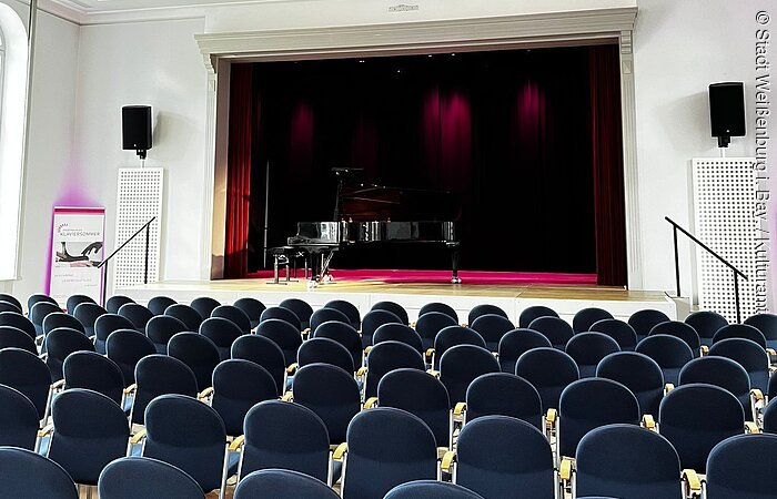 Leerer Konzertsaal mit Reihen blauer Stühle und einem Flügel auf der Bühne unter einer Deckenleuchte.