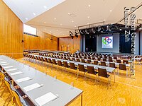 Saal der Stadthalle Gunzenhausen Großer Konferenzraum mit Reihen von Tischen und Stühlen, Bühne mit Rednerpult und Leinwand im Hintergrund.