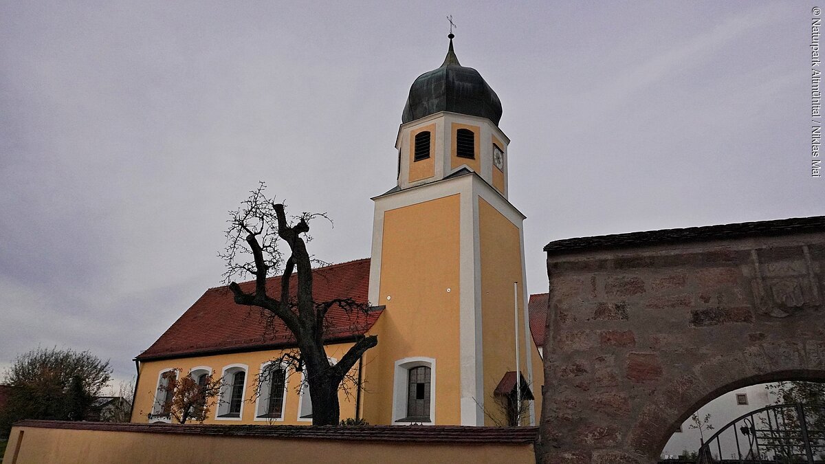 Gunzenhausen Kirche Cronheim Außenansicht Kirchturm mit Zwiebeldach und gelbes Kirchengebäude hinter einer Mauer bei bewölktem Himmel.