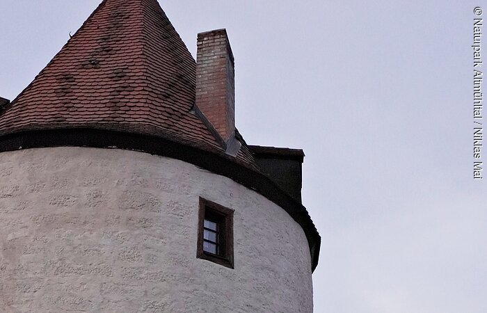 Runder weißer Turm mit roten Dachziegeln, zwei kleinen Fenstern und Laterne an der Wand bei Dämmerung.