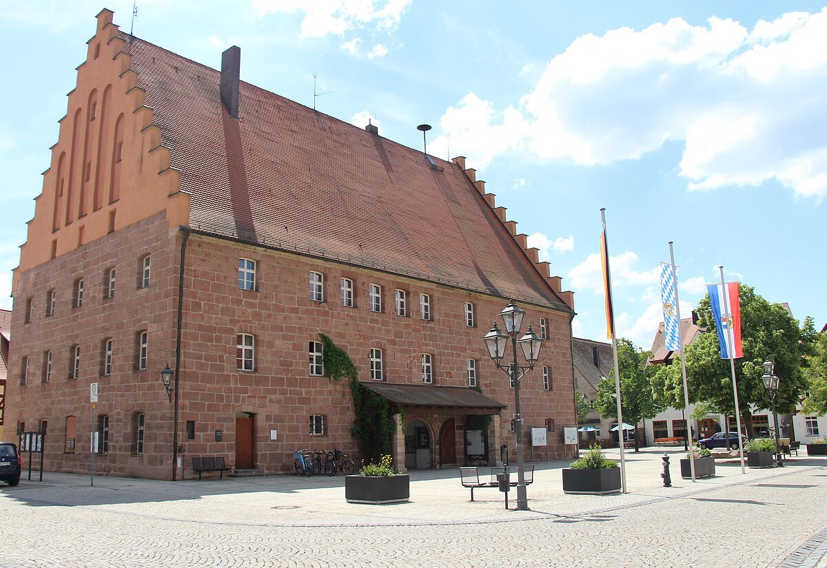 Großes historisches Backsteingebäude mit Treppengiebel und drei Flaggen auf einem gepflasterten Platz bei Tageslicht.