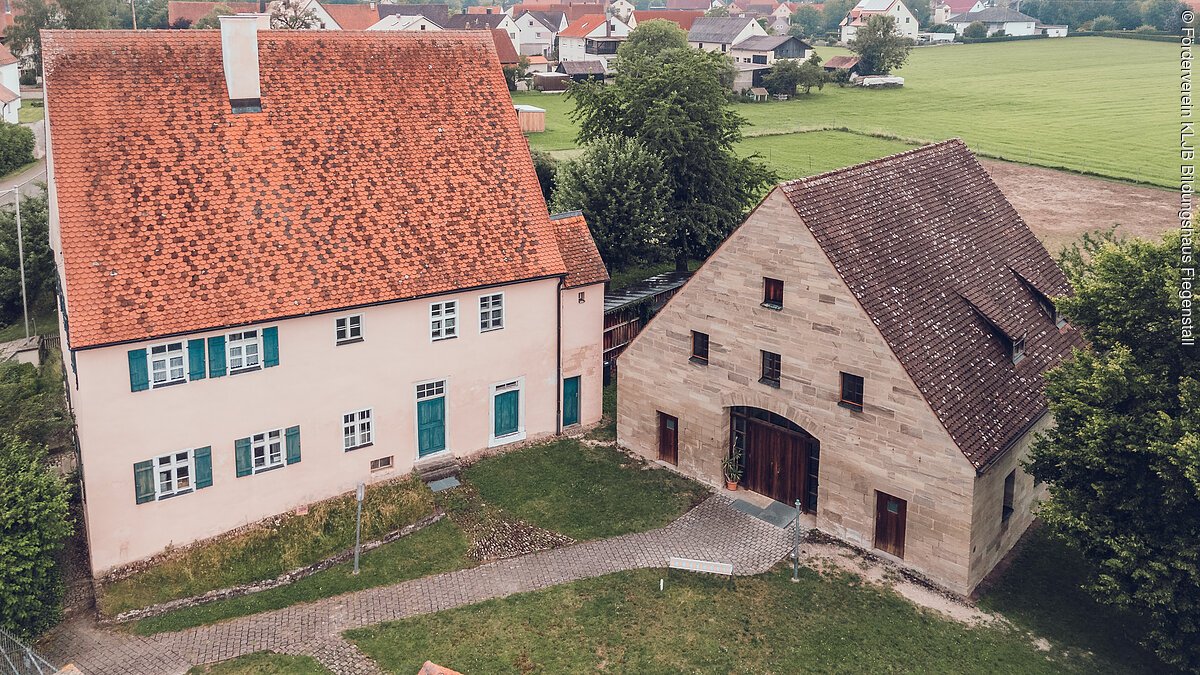 KLJB Bildungshaus Fiegenstall Luftaufnahme von zwei Gebäuden mit roten Dächern und grünem Rasen dazwischen in einem Dorf.